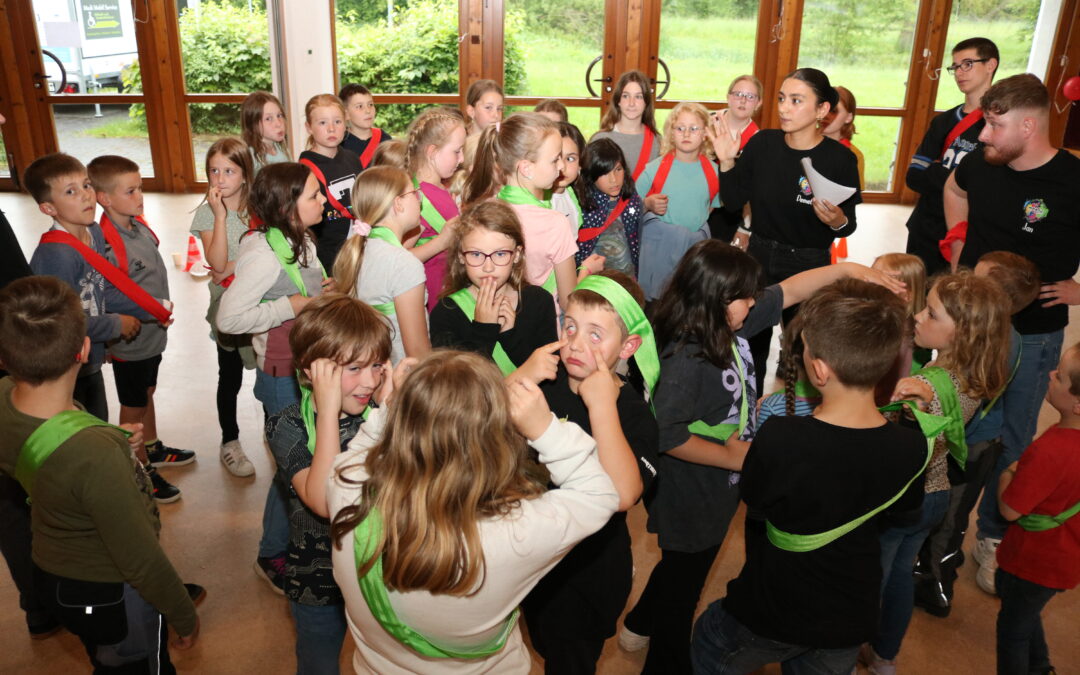 Gruppe von Kindern in einem Raum, einige tragen grüne und rote Bänder. Sie stehen in einem Kreis und scheinen miteinander zu kommunizieren.