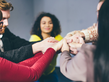 Mehrere Hände, die übereinander gestapelt sind, während eine Person im Hintergrund lächelt. Teamarbeit und Zusammenarbeit werden symbolisiert.
