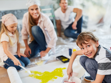 Fröhliche Familie sitzt während Renovierungsarbeiten auf dem Boden, knieendes Mädchen mit Kopftuch malt mit Pinsel auf dem Boden liegende Tapete gelb an, Junge hockt daneben und lacht
